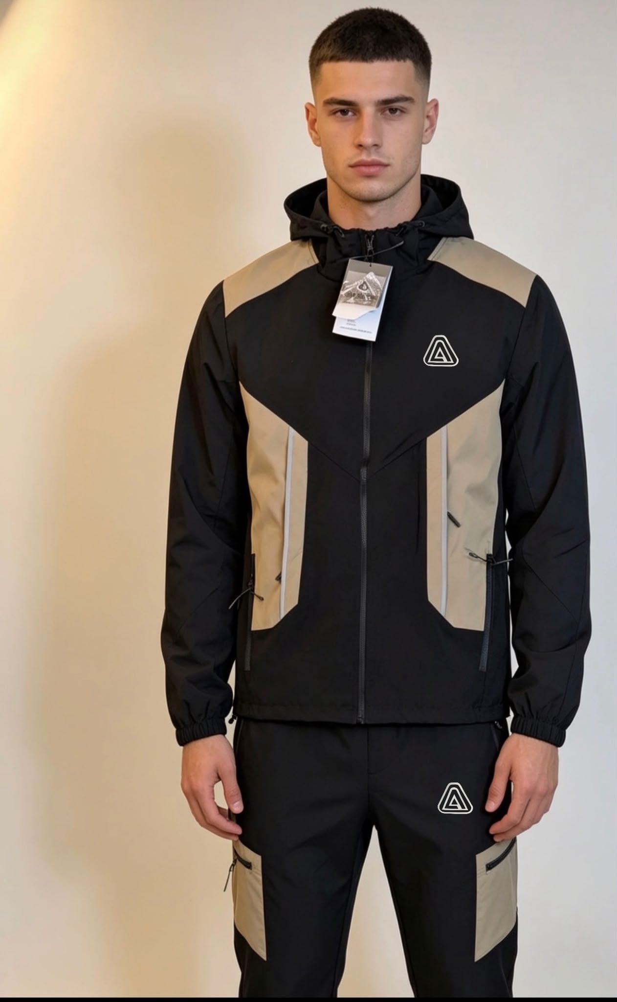 A man modeling a black and beige hooded jacket with matching black pants, featuring a small white geometric logo on the chest and thigh.