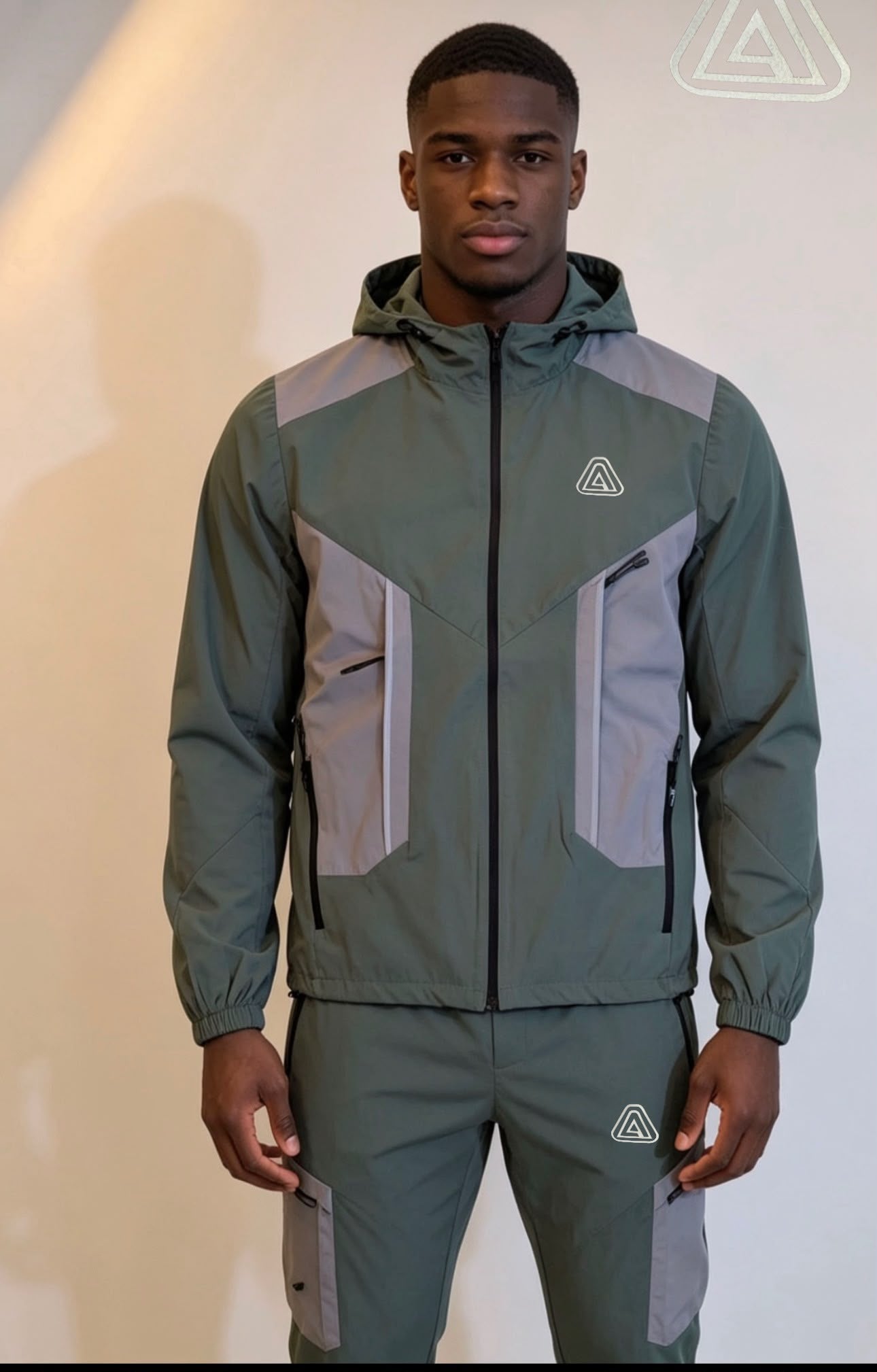 A man modeling a matching hooded jacket and pants set in muted sage green and light gray, featuring a small white geometric logo on the chest and thigh.
