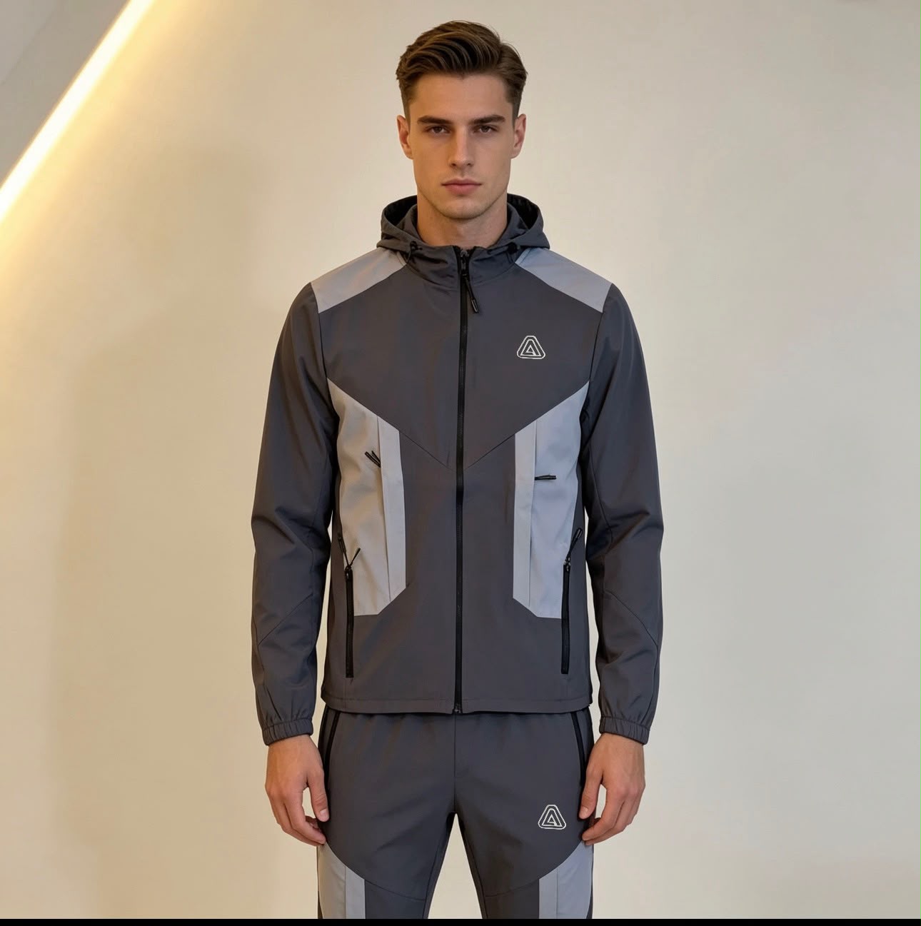A man modeling a dark gray and light gray athletic windbreaker jacket with matching joggers, featuring a small geometric logo on the chest and thigh.